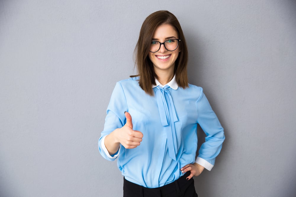Happy businesswoman showing thumb up over gray background. Wearing in blue shirt and glasses. Looking at camera Happy businesswoman showing thumb up over gray background. Wearing in blue shirt and glasses. Looking at camera